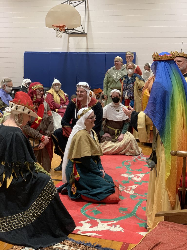 Baroness Sciath kneels in front of Queen Kayla, surrounded by the order of the Pelican.