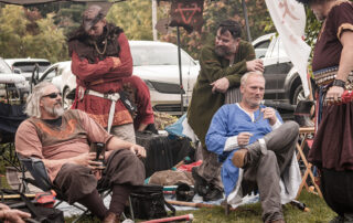 A group of people dressed in tunics sit in chairs outside in a field beneath a sunshade at Huntsman's Harvest, smiling and chatting