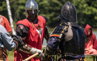 Sir Maneke dressed in a red tabard uses a polearm to fight incoming Barbarian fighters
