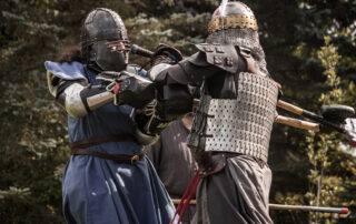 Hespeler in a feathered helmet and lamellar armour and Vivian, dressed in blue with a dark steel helmet, fight up-close during a tournament