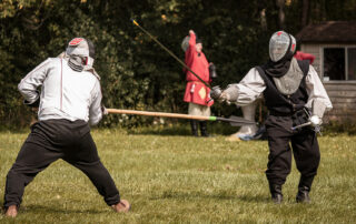 A fencer dressed in white, wielding a rattan spear, throws a thrust against another fencer dressed in black, fighting with two silver swords