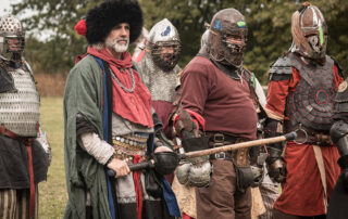 Noble Hespeler wearing a large fuzzy hat and multiple layers of clothes stands with the armoured fighters