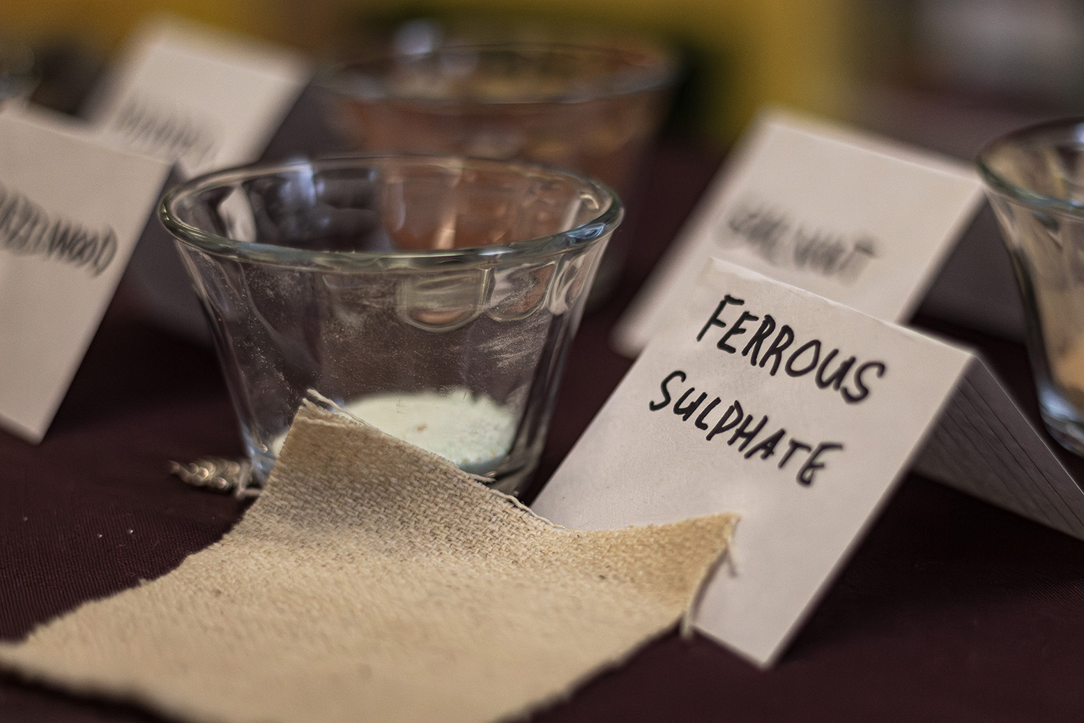 A bolt of tan fabric against a glass container full of powder and a paper card labeled Ferrous Sulphate