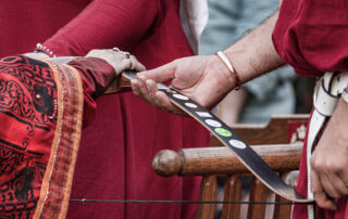 Nika's hand holds the top of a bow adorned with stickers. Their Majesties' hands gently hold the underside of the same bow as Nika swears fealty